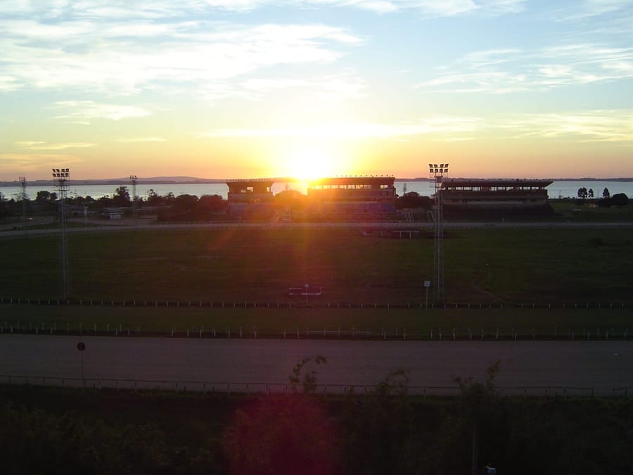 Pôr do sol sobre o Guaíba visto por trás das tribunas do Hipódromo do Cristal, com a pista em primeiro plano